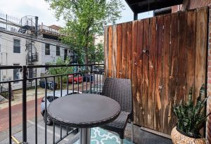 Small balcony with a round table, chair, and potted plant, overlooking a street with buildings and a tree.